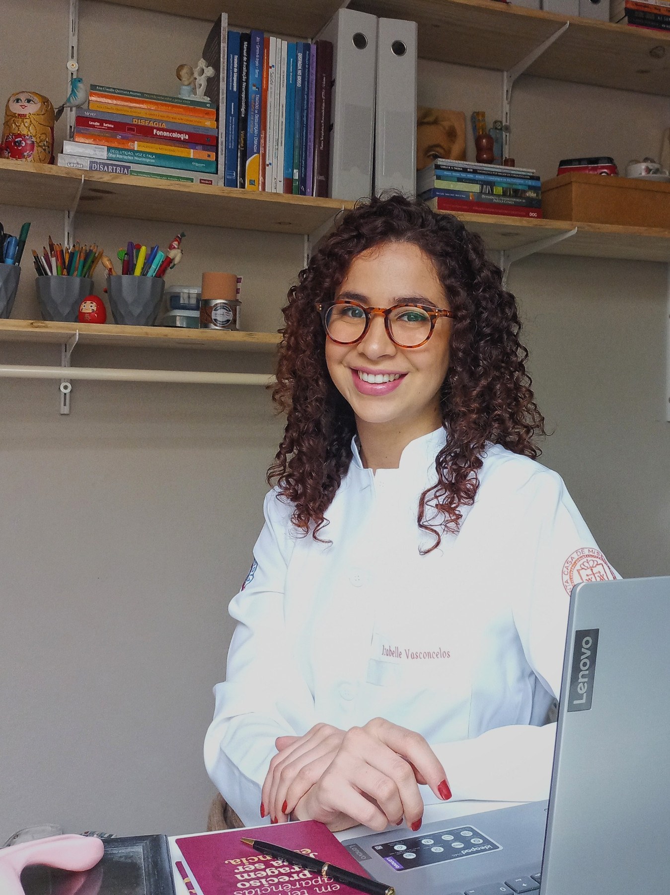 Izabelle Vasconcelos de jaleco, sorrindo, em frente a uma estante